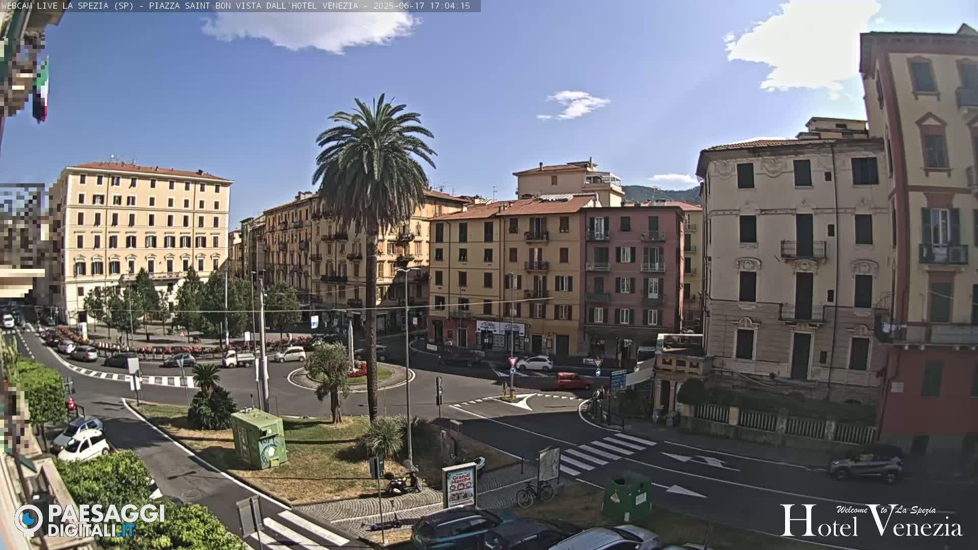 A sunny day in a European city square, featuring several multi-story buildings, a roundabout with cars, and a tall palm tree in the center.