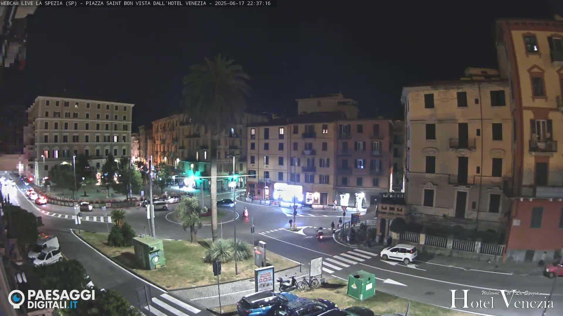 A nighttime view of a city square with several multi-story buildings, some cars, and a palm tree, under a clear sky.
