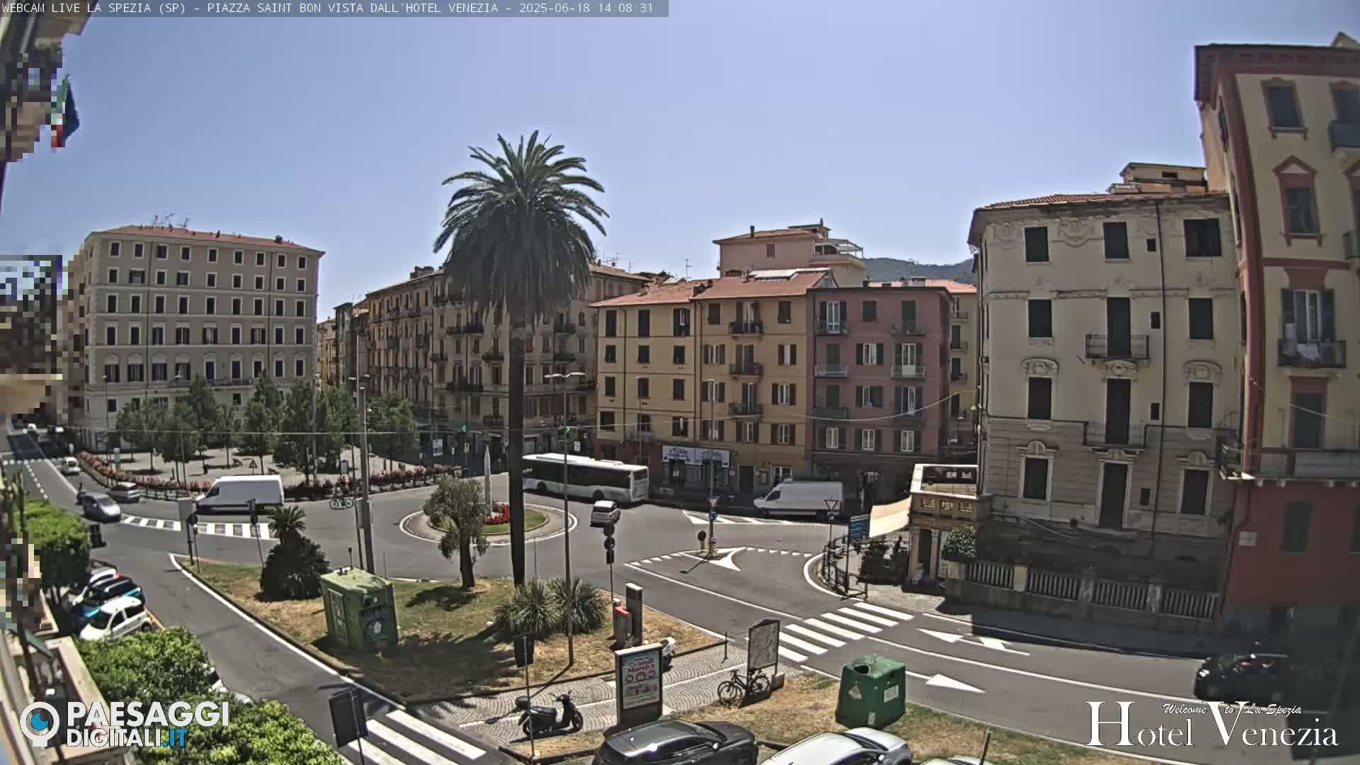 A sunny city square in Italy, featuring several multi-story buildings, a palm tree, vehicles, and pedestrian crossings.