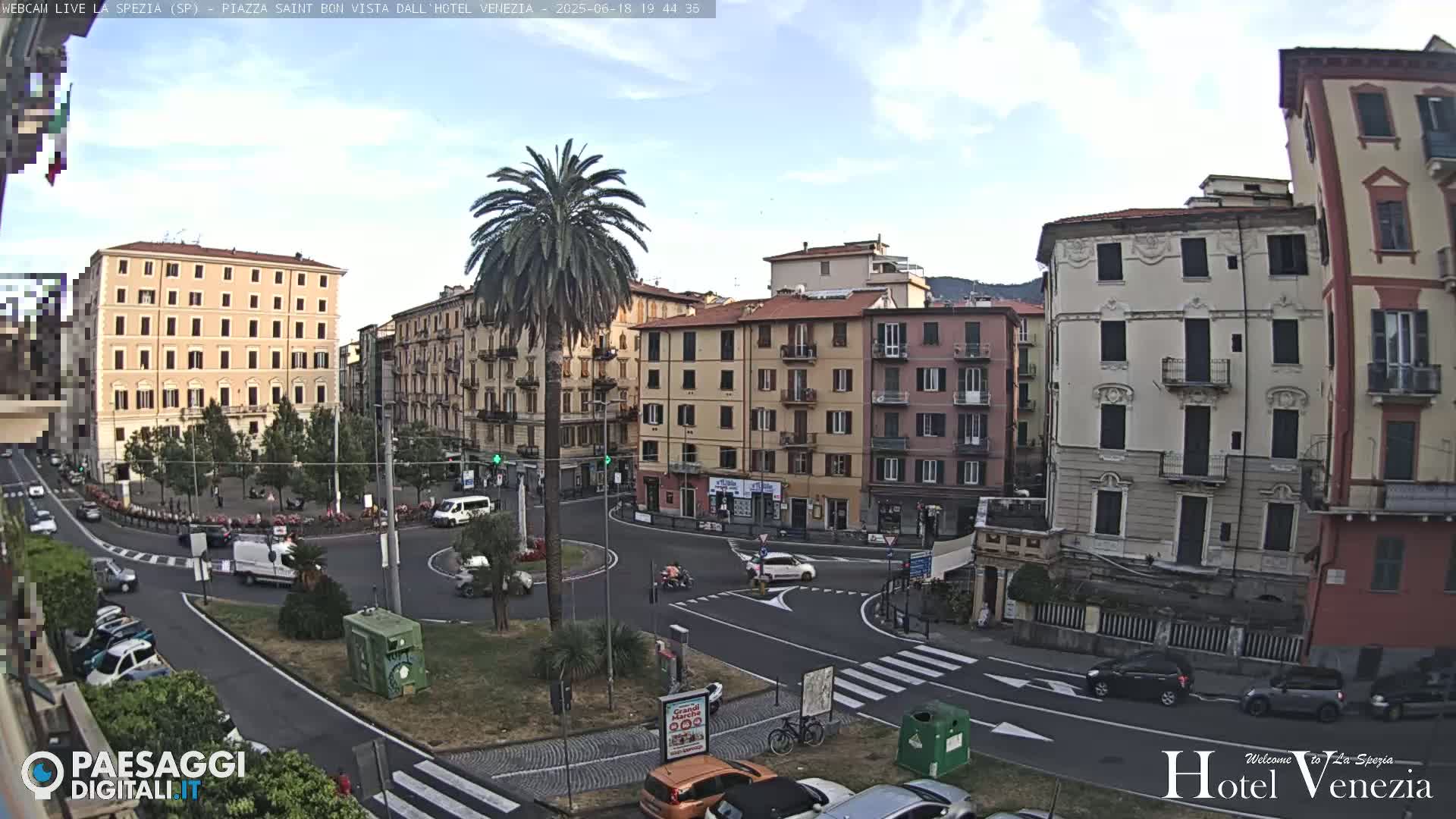 A mostly sunny view of a European city square with several multi-story buildings surrounding a roundabout where cars are driving.