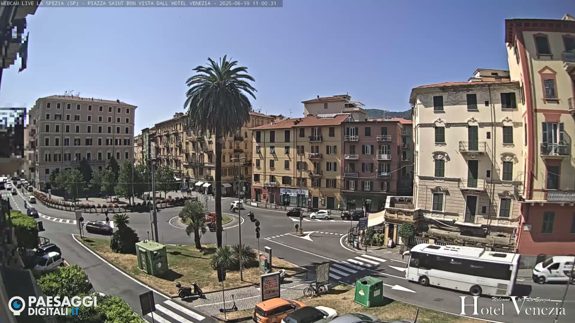 A sunny day in a European city square with several multi-story buildings surrounding a circular road, and a palm tree in the center.