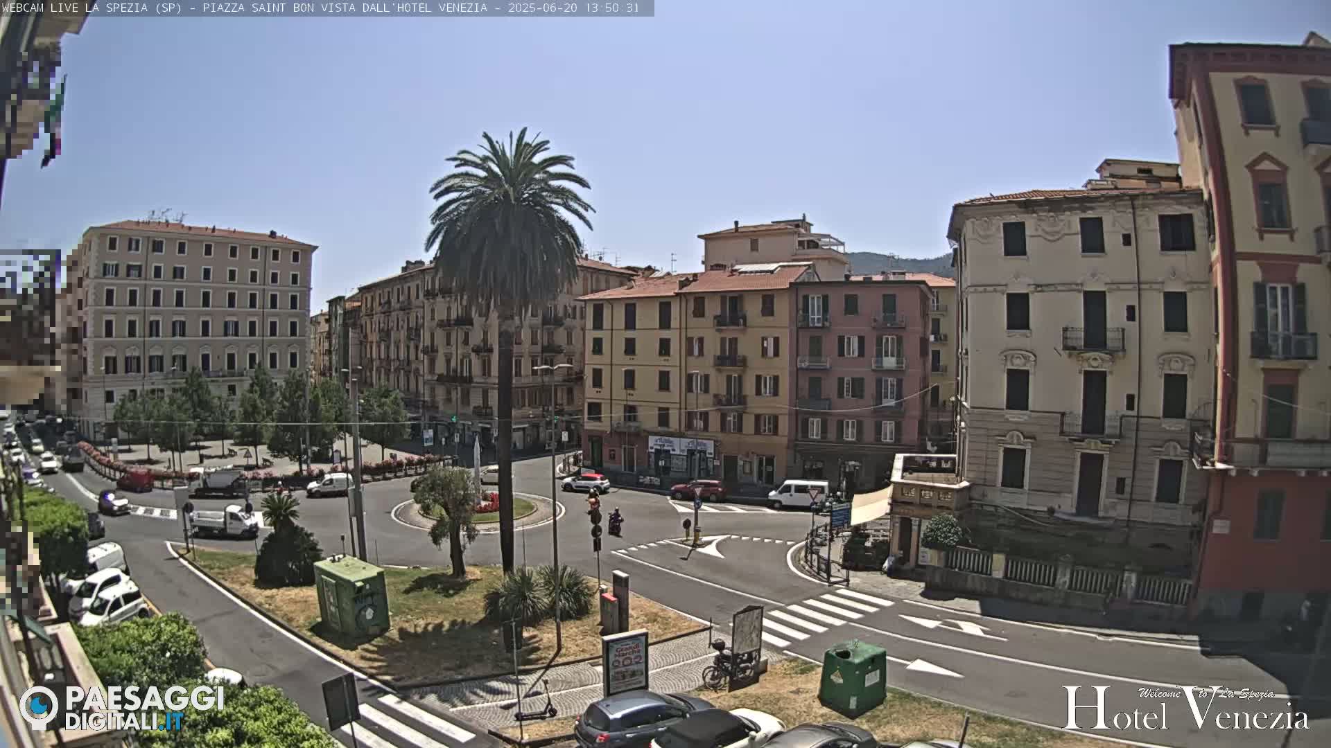 A sunny day reveals a city square surrounded by multi-story buildings, with vehicles and pedestrians navigating a roundabout.