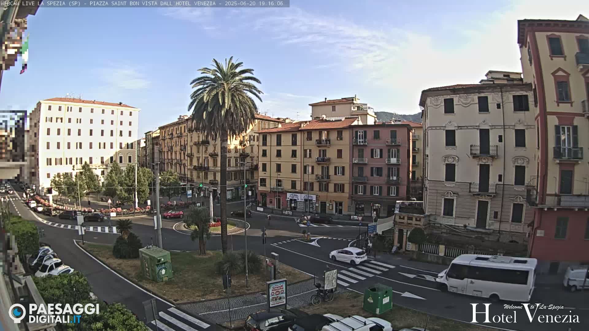 A sunny day reveals a city square with multi-story buildings, a palm tree in the center, and cars driving around the roundabout.