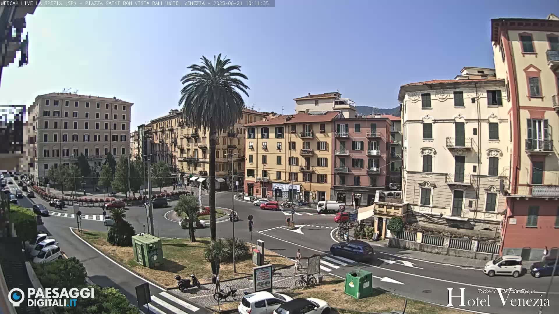 A sunny day in a European city square with several multi-story buildings surrounding a roundabout with cars and pedestrians.