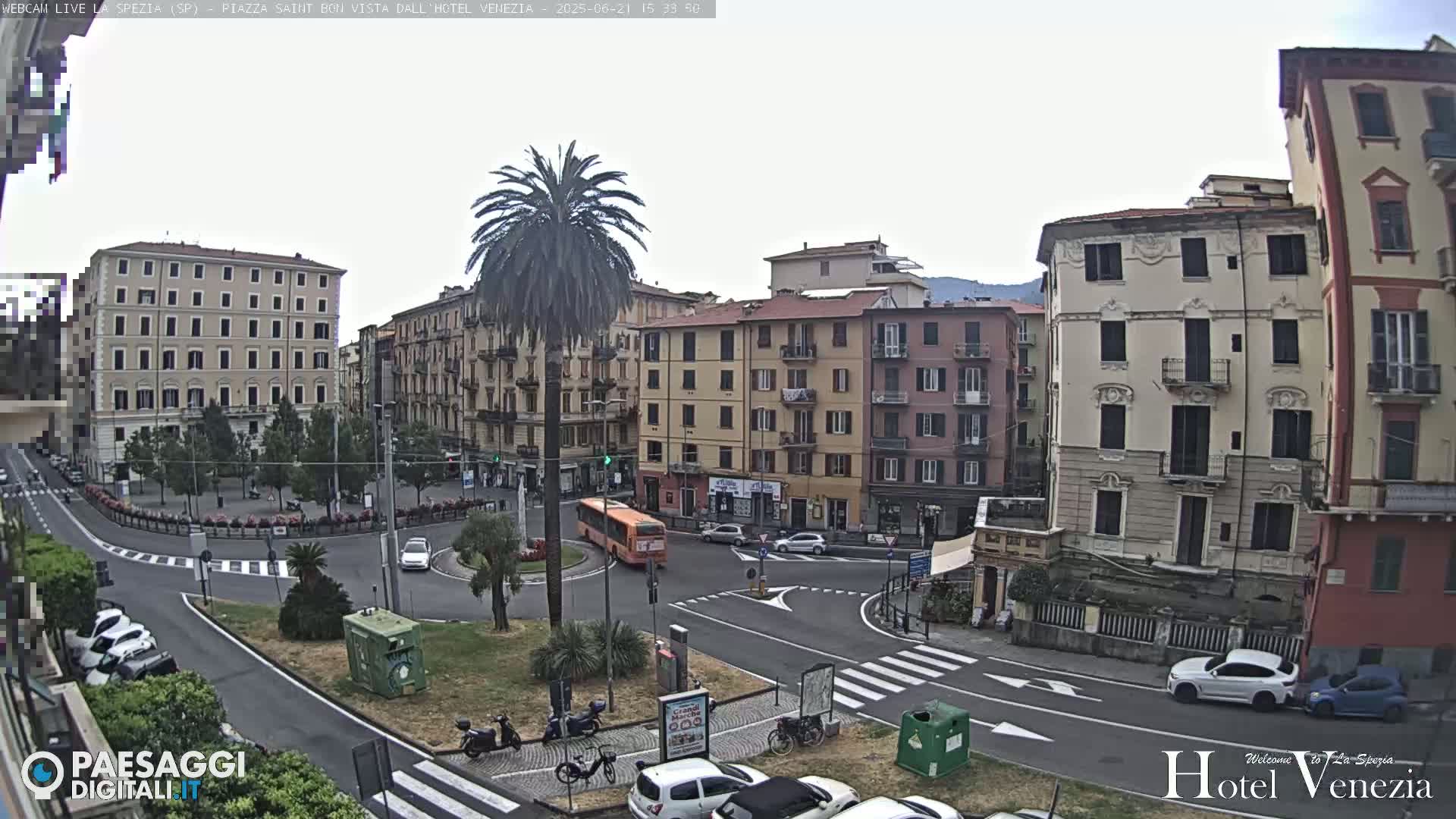 A mostly sunny day in a European city square with several multi-story buildings, a palm tree in the center, cars parked along the sides of the streets, and an orange bus traveling through a roundabout.