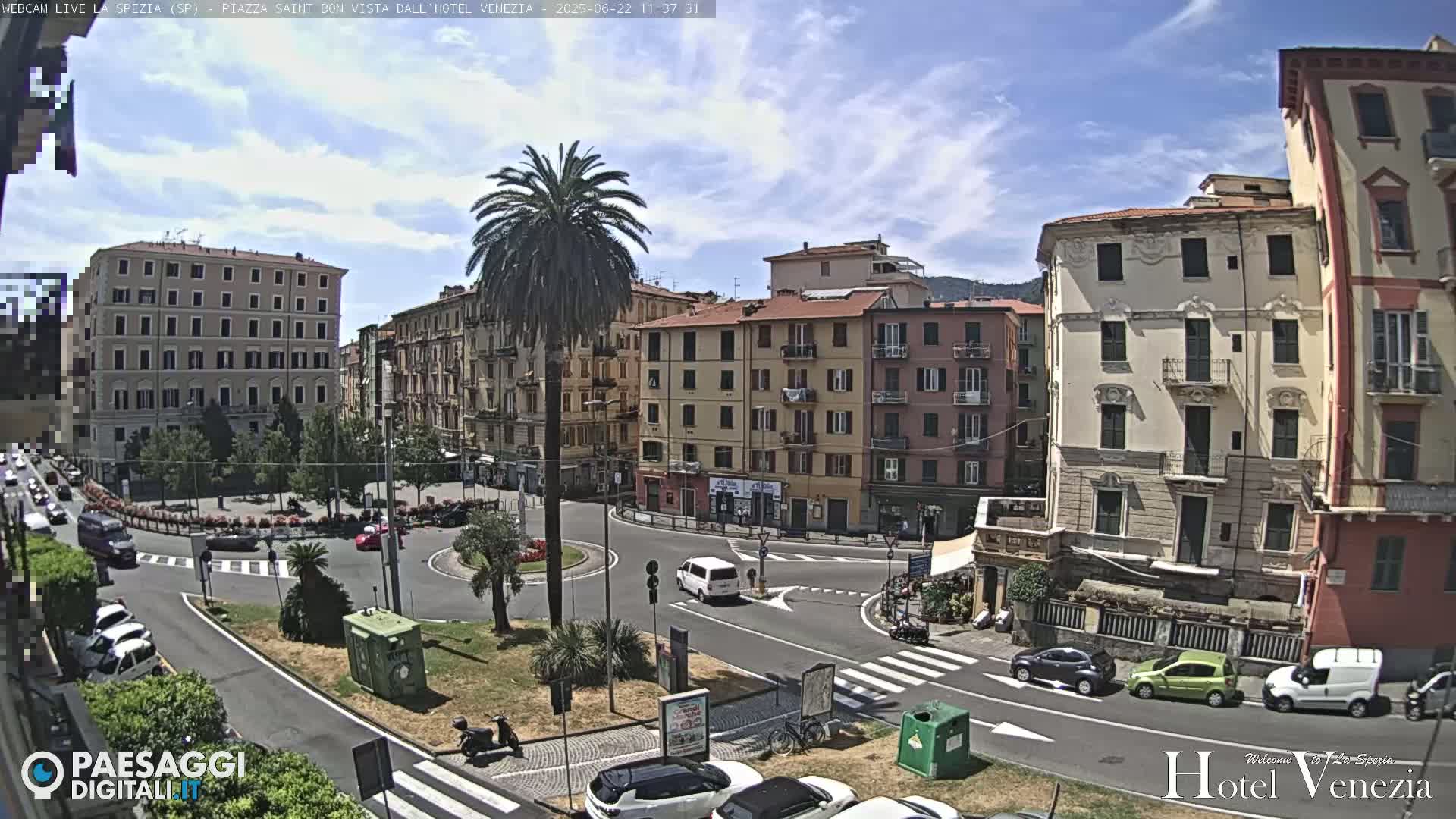 A sunny day in a European city square, showing several multi-story buildings surrounding a roundabout with cars and a few pedestrians.