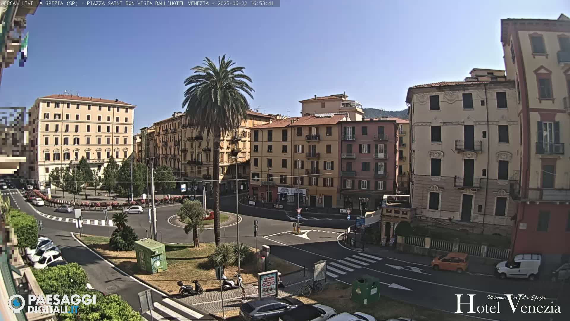 A sunny day in a European city square, showing several multi-story buildings surrounding a traffic circle with parked cars and a few pedestrians.