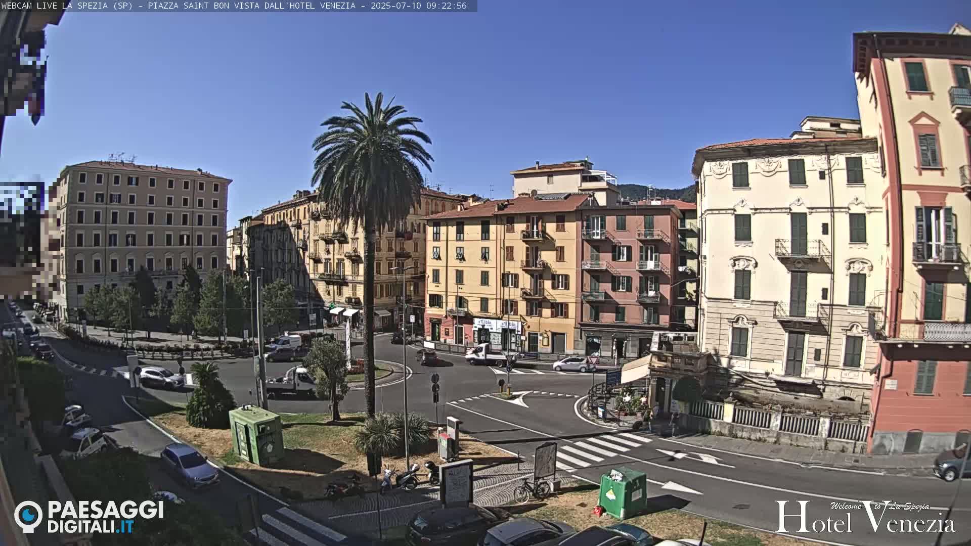A sunny day in a European city square shows several multi-story buildings surrounding a roundabout with cars and a palm tree in the center.