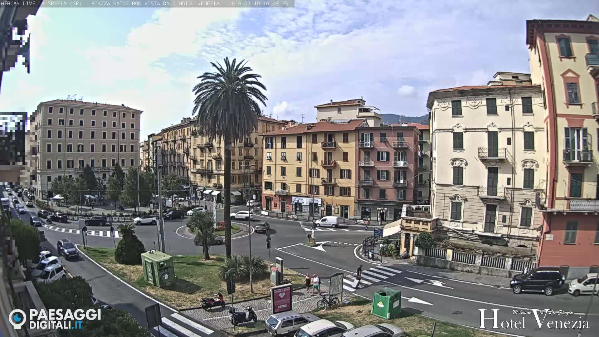 A sunny day in a European city square shows several multi-story buildings surrounding a roundabout with cars and pedestrians.