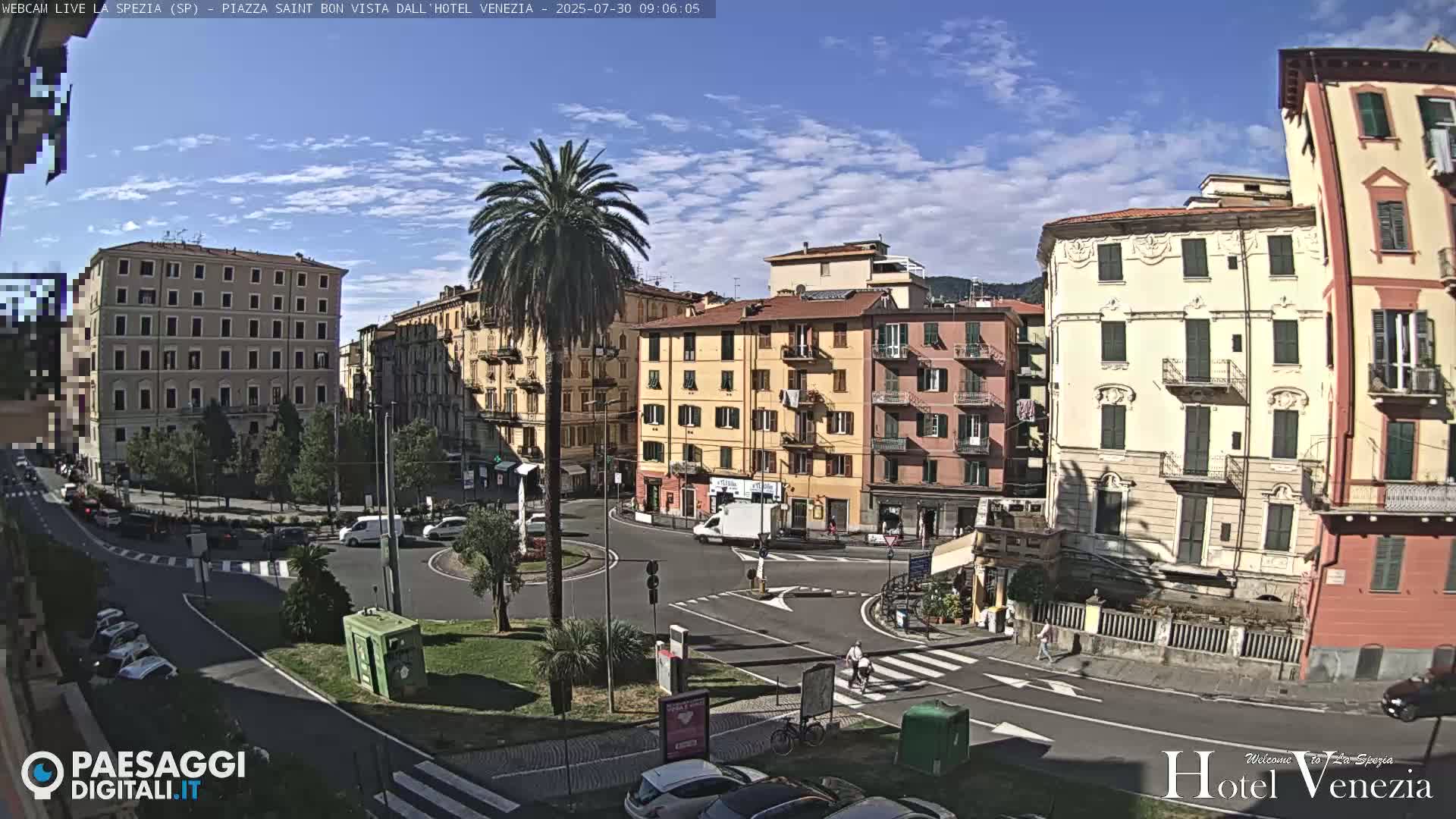 A sunny day in a European city square shows several multi-story buildings surrounding a roundabout with cars and pedestrians.