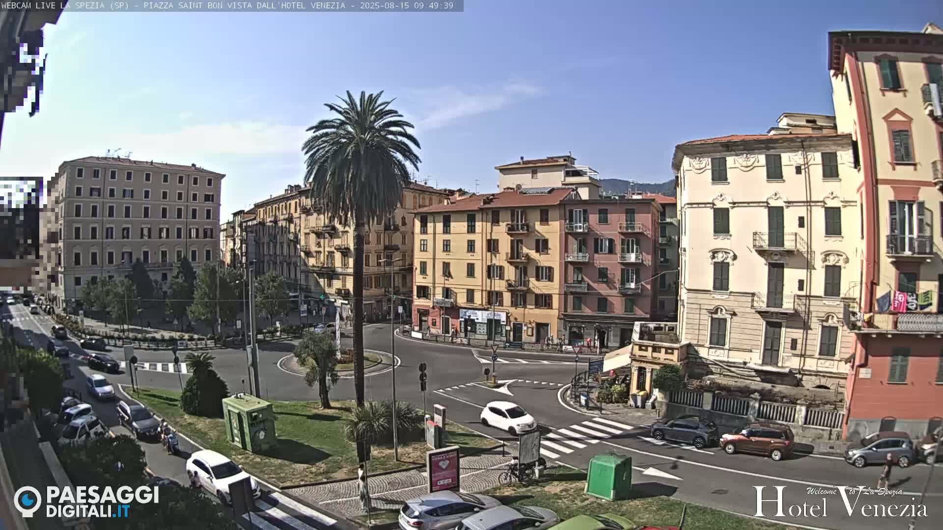 A sunny day reveals a circular plaza in a city, surrounded by multi-story buildings, with cars and a palm tree visible.