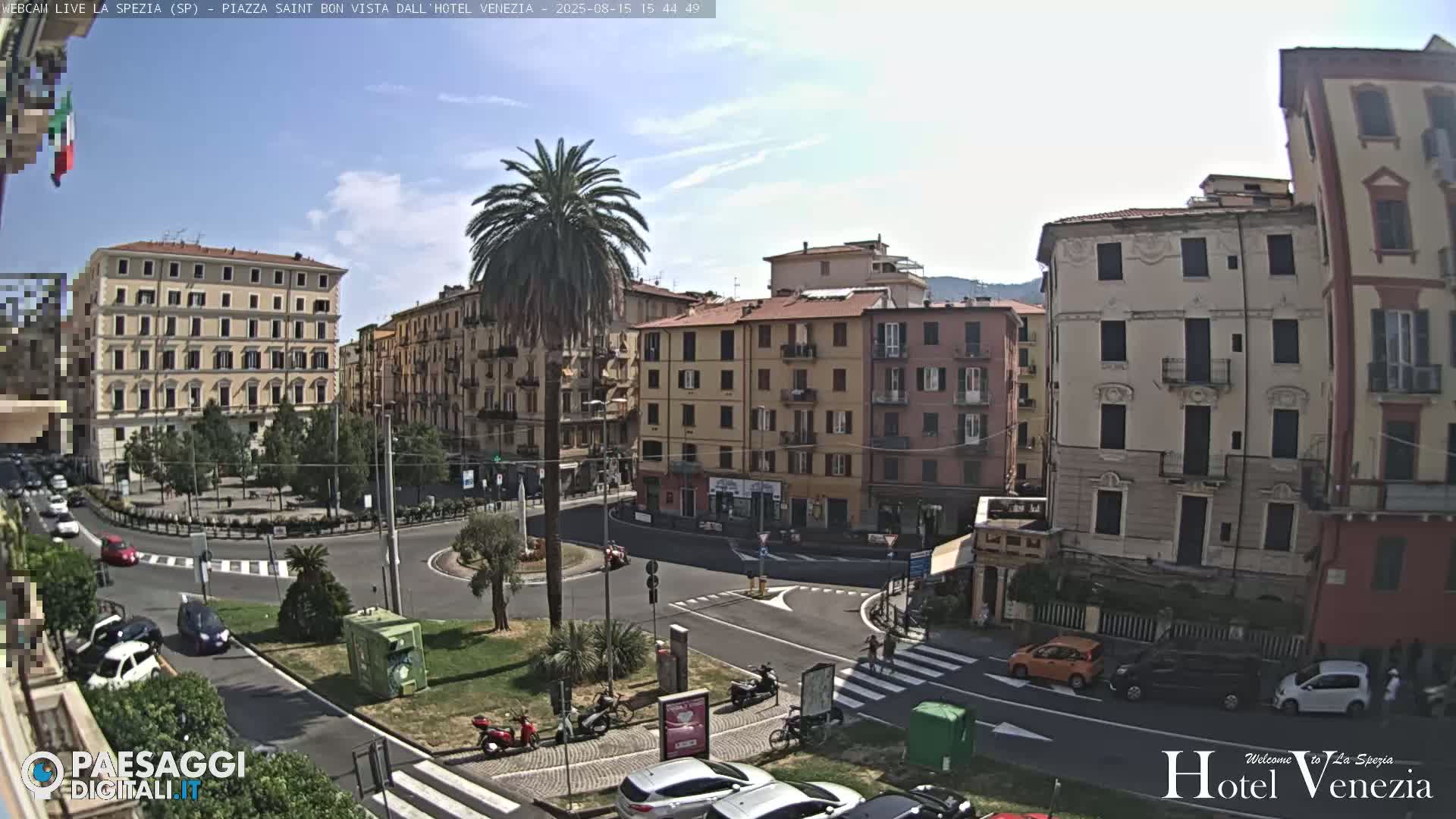 A sunny day reveals a circular plaza in a city, surrounded by multi-story buildings, with cars and a palm tree visible.