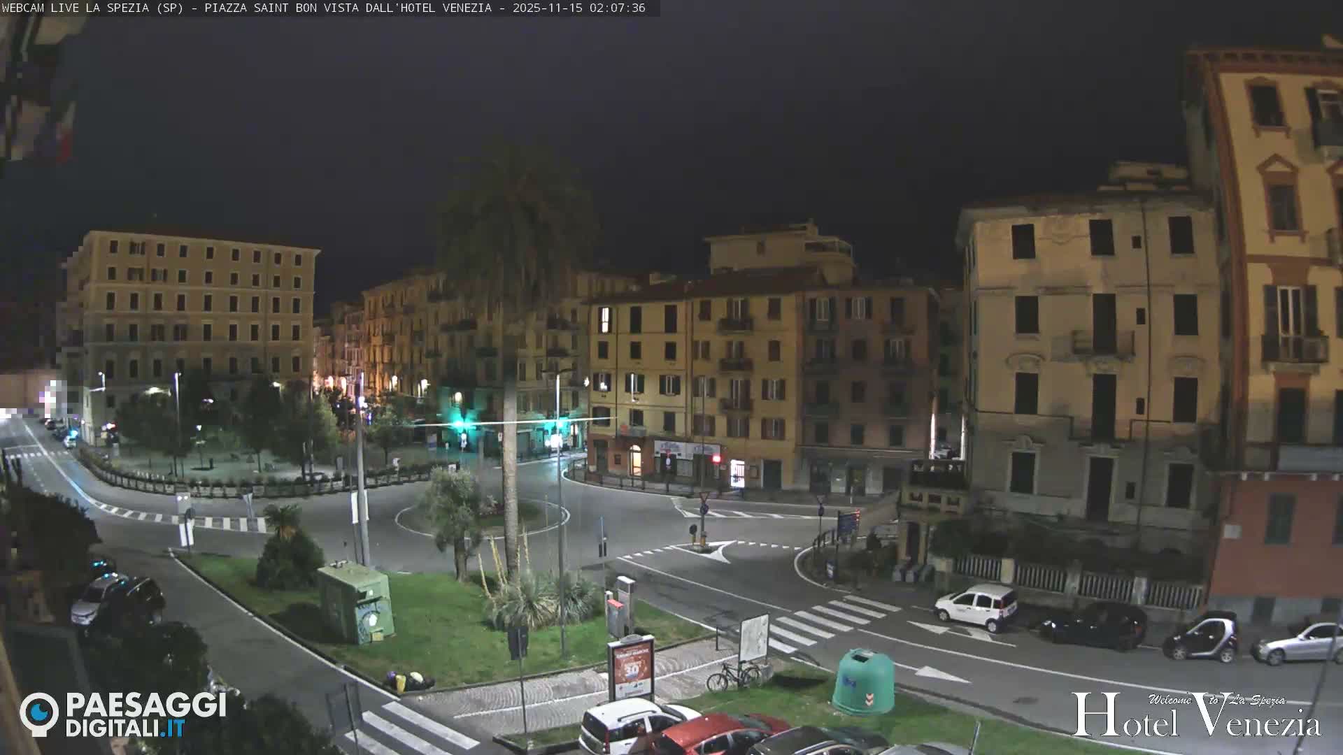 An illuminated urban street scene at night depicts a wide roundabout surrounded by multi-story buildings with some lit windows, parked cars, and scattered trees, all under a clear, dark sky.
