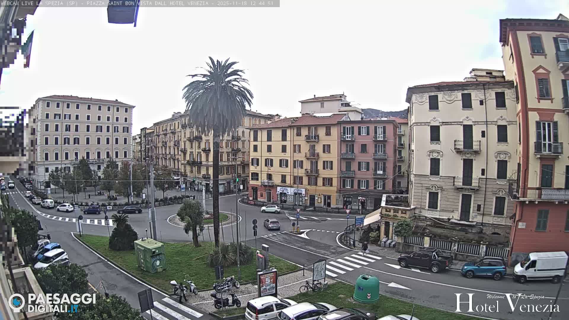 An illuminated urban street scene at night depicts a wide roundabout surrounded by multi-story buildings with some lit windows, parked cars, and scattered trees, all under a clear, dark sky.