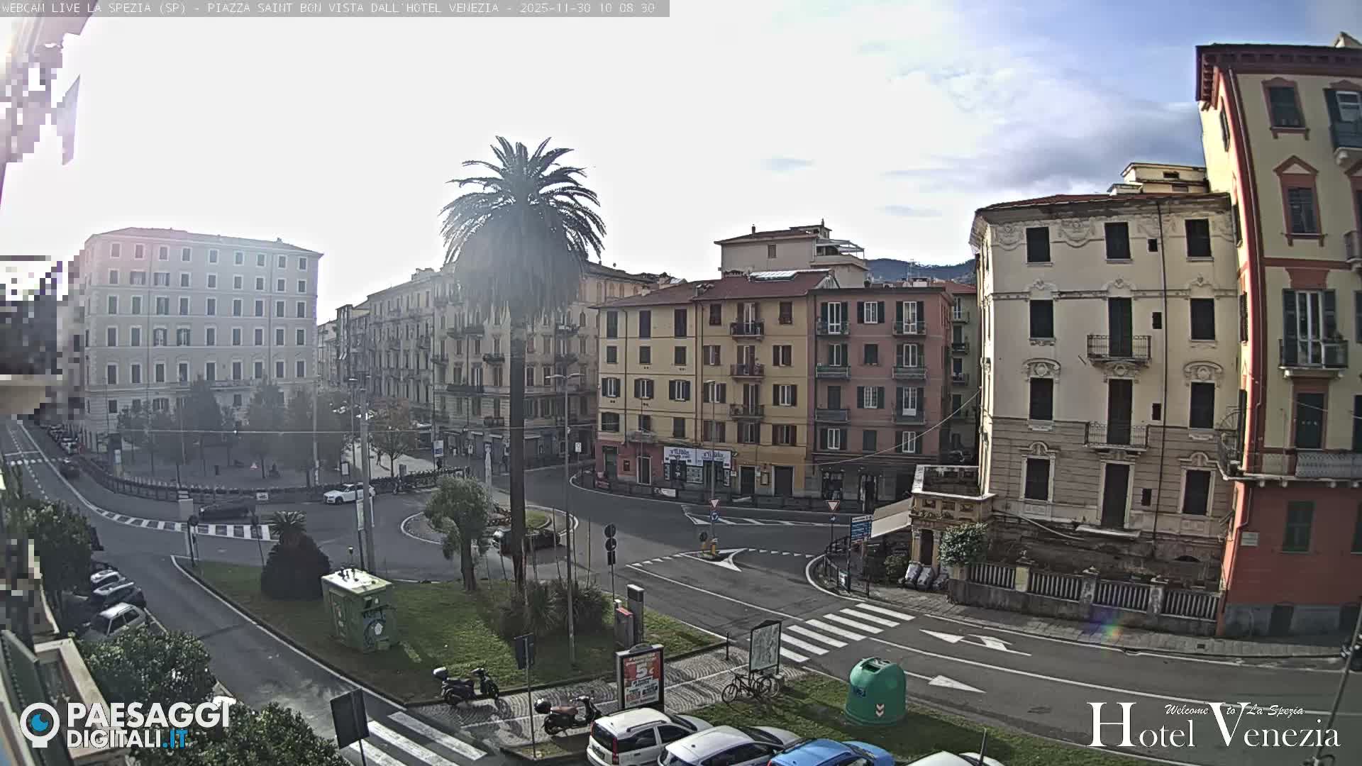 An urban square is visible with a prominent palm tree, a roundabout bustling with cars and scooters, and numerous multi-story buildings in a variety of pastel colors, all under a partly cloudy sky.