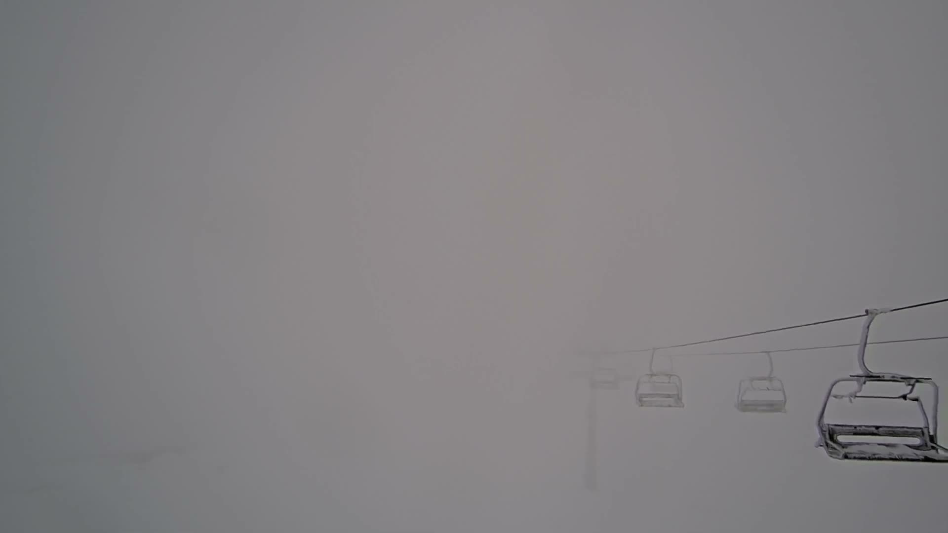 Empty ski lift chairs, some lightly frosted, disappear into a thick, gray fog, indicating extremely low visibility and cold, overcast weather.