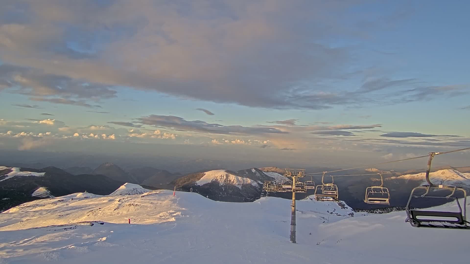 A snow-covered mountain ski slope featuring a prominent ski lift is visible under a partly cloudy sky, illuminated by warm, golden light.