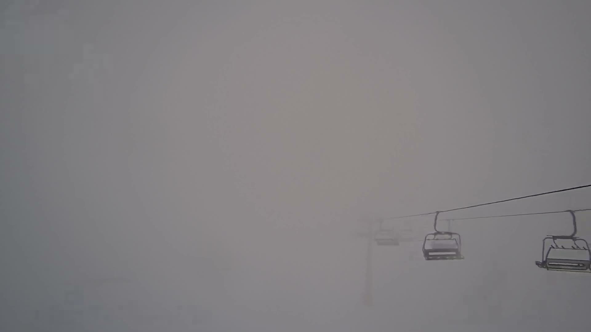 Several empty chairlift chairs and a support tower are partially visible through extremely dense fog or heavy clouds at a ski resort.