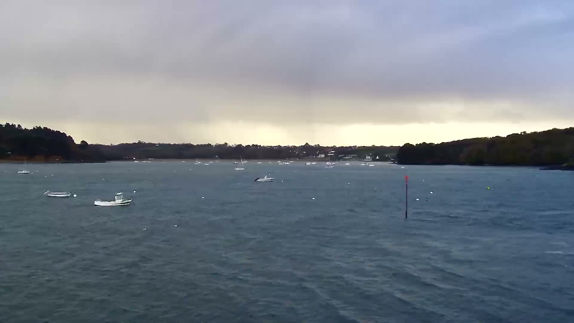 Numerous small boats are moored across a choppy bay under an overcast sky with a lighter horizon, flanked by a dark, tree-lined coast dotted with distant buildings.