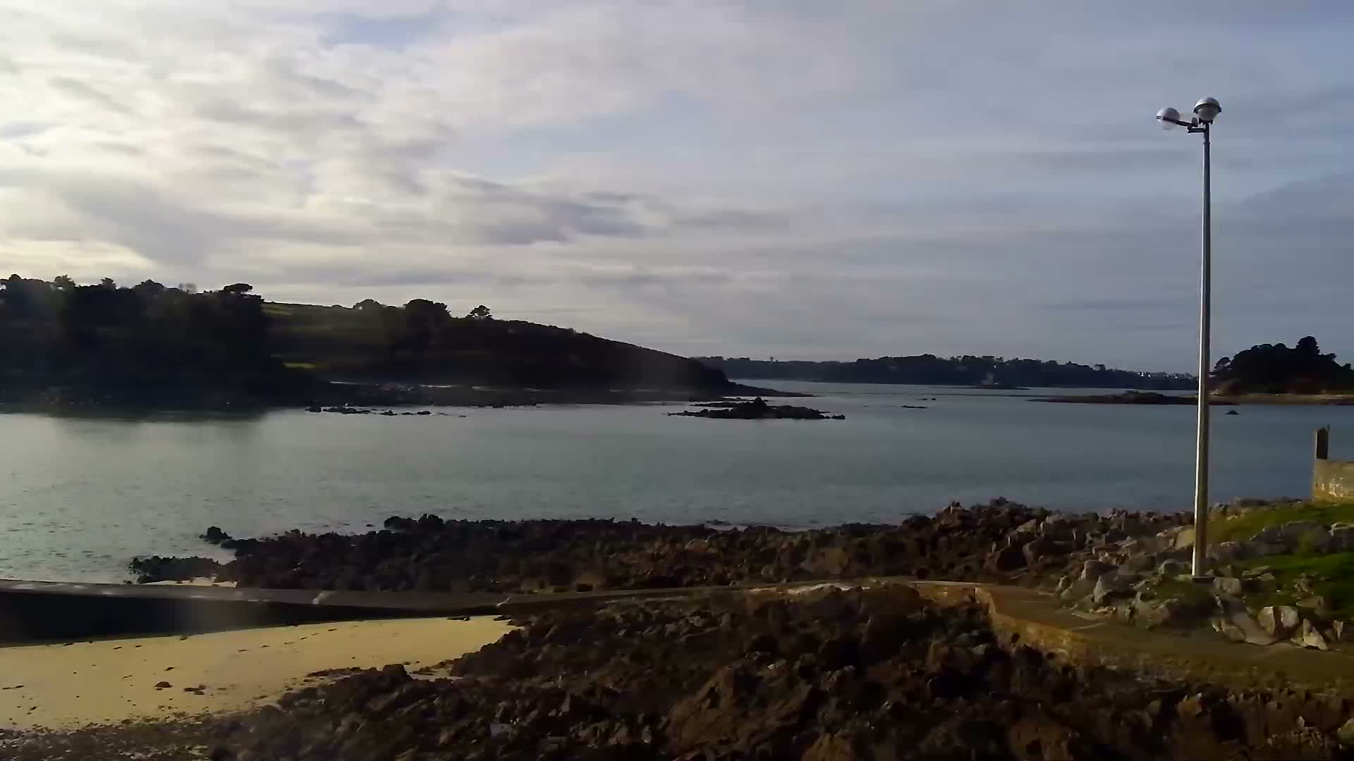 Numerous small boats are moored across a choppy bay under an overcast sky with a lighter horizon, flanked by a dark, tree-lined coast dotted with distant buildings.