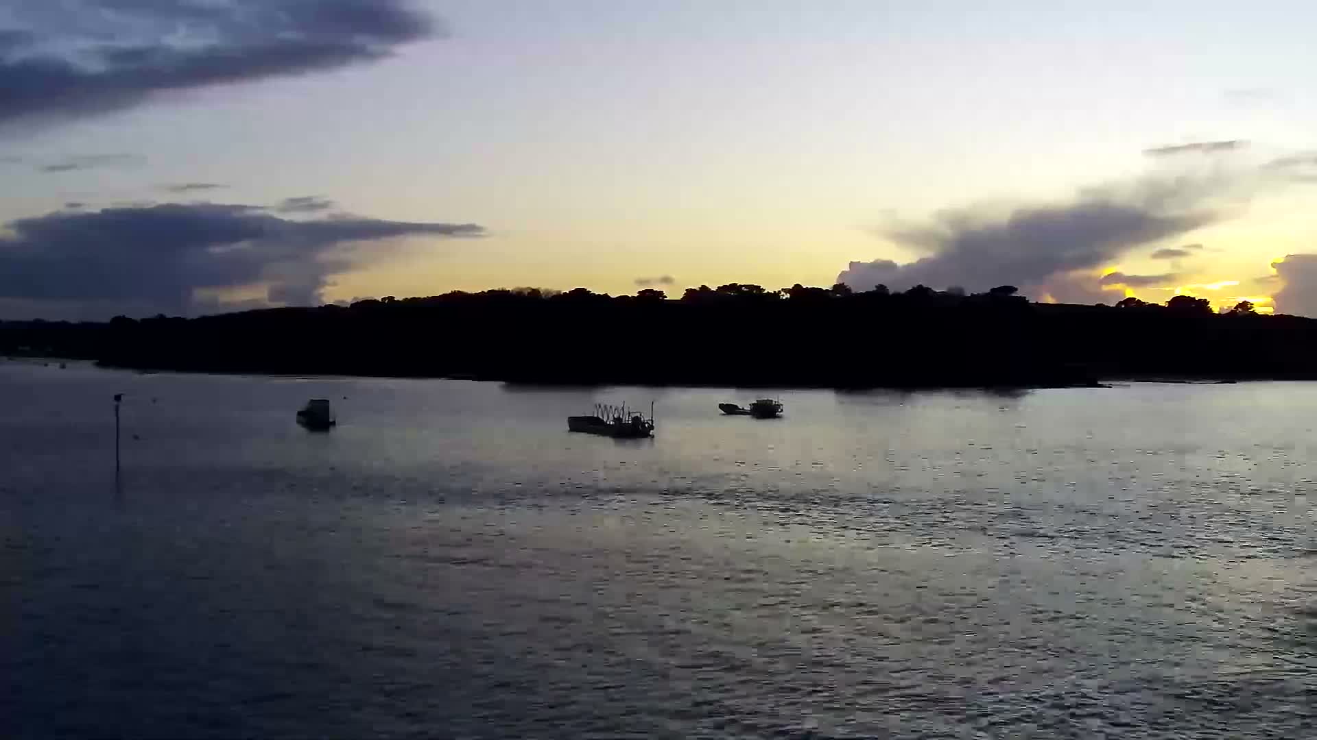 Boats dot the dark, calm water of a river or bay, with a distant treeline silhouetted against a cloudy sky featuring vibrant orange and yellow hues of either sunrise or sunset.