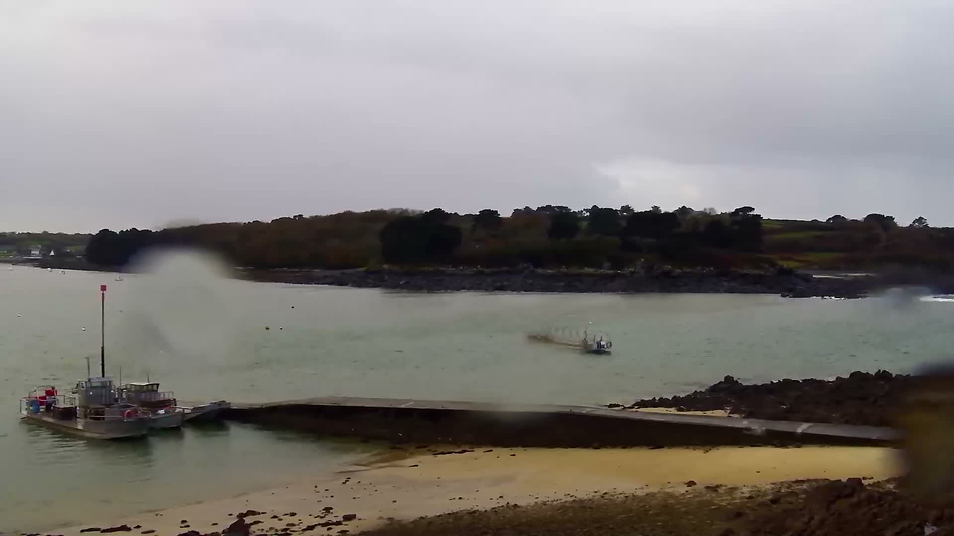 Under an overcast and possibly rainy sky, two utility boats are docked at a concrete pier on a sandy and rocky shore within a murky green bay, with a smaller vessel visible further out and a dark, tree-lined coast with distant houses in the background.