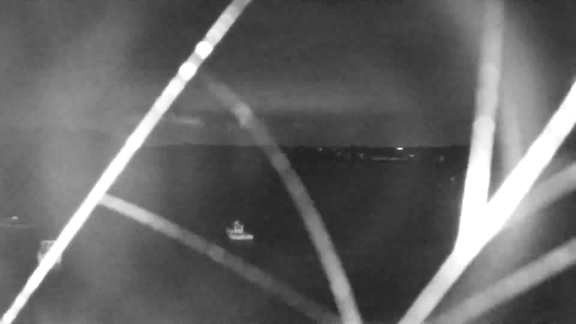 Under an overcast and possibly rainy sky, two utility boats are docked at a concrete pier on a sandy and rocky shore within a murky green bay, with a smaller vessel visible further out and a dark, tree-lined coast with distant houses in the background.