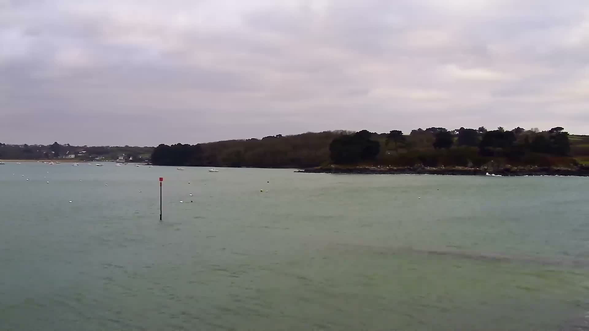 Under an overcast and possibly rainy sky, two utility boats are docked at a concrete pier on a sandy and rocky shore within a murky green bay, with a smaller vessel visible further out and a dark, tree-lined coast with distant houses in the background.