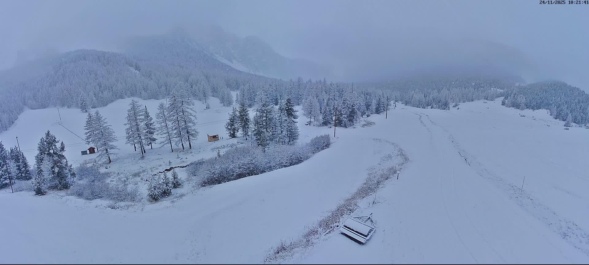 A vast, snow-covered mountain landscape featuring dense forests, ski lift poles, a small cabin, and a piece of snow grooming equipment in the foreground is visible under an overcast and foggy sky.