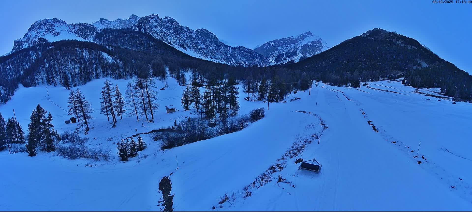A snow-covered mountain valley features a ski slope, scattered trees, and distant snow-capped peaks under a clear, cold twilight sky.