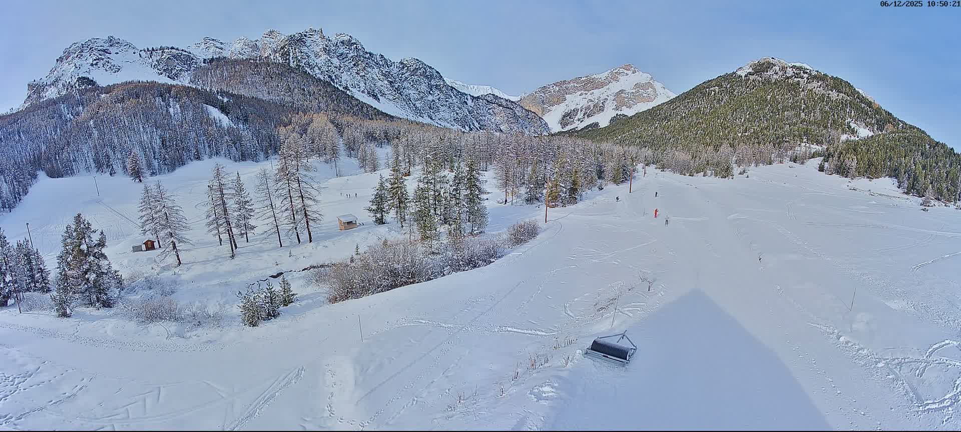 This image depicts a vast, snow-covered mountain landscape with a ski slope winding through snow-dusted evergreen and bare deciduous trees, featuring several skiers under a clear blue sky.