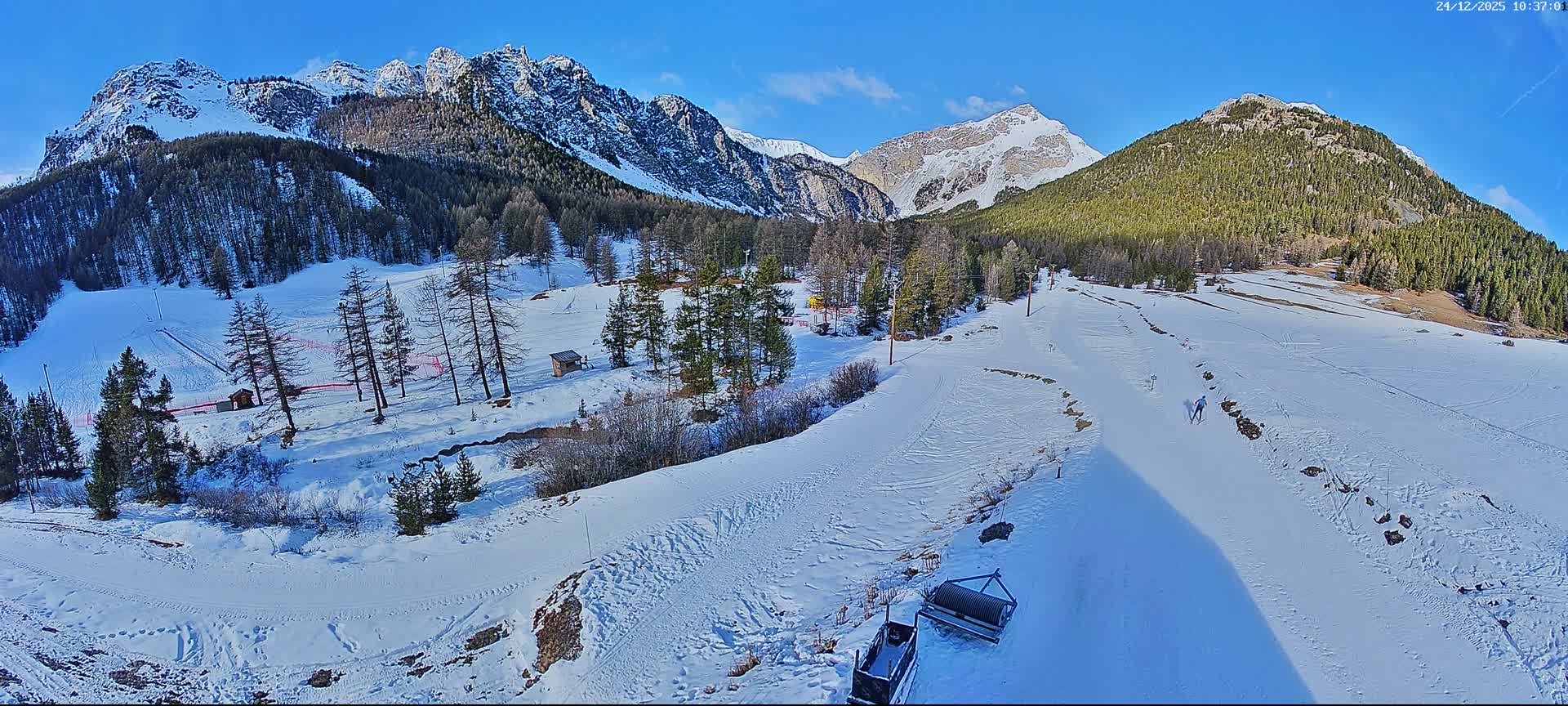 This image depicts a vast, snow-covered mountain landscape with a ski slope winding through snow-dusted evergreen and bare deciduous trees, featuring several skiers under a clear blue sky.