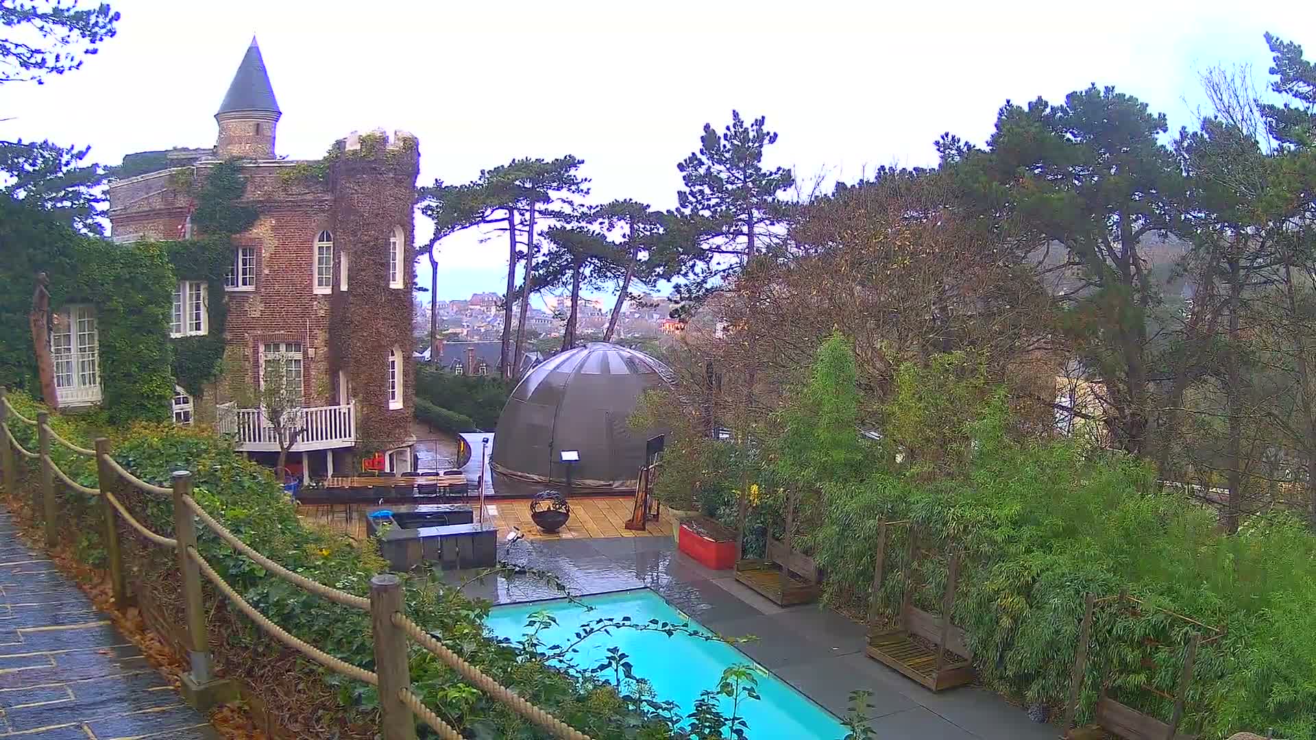 Under an overcast sky, a vine-covered brick building with a turret overlooks a modern dome, a bright blue swimming pool, and a wet stone path, all nestled among dense trees and a distant town.