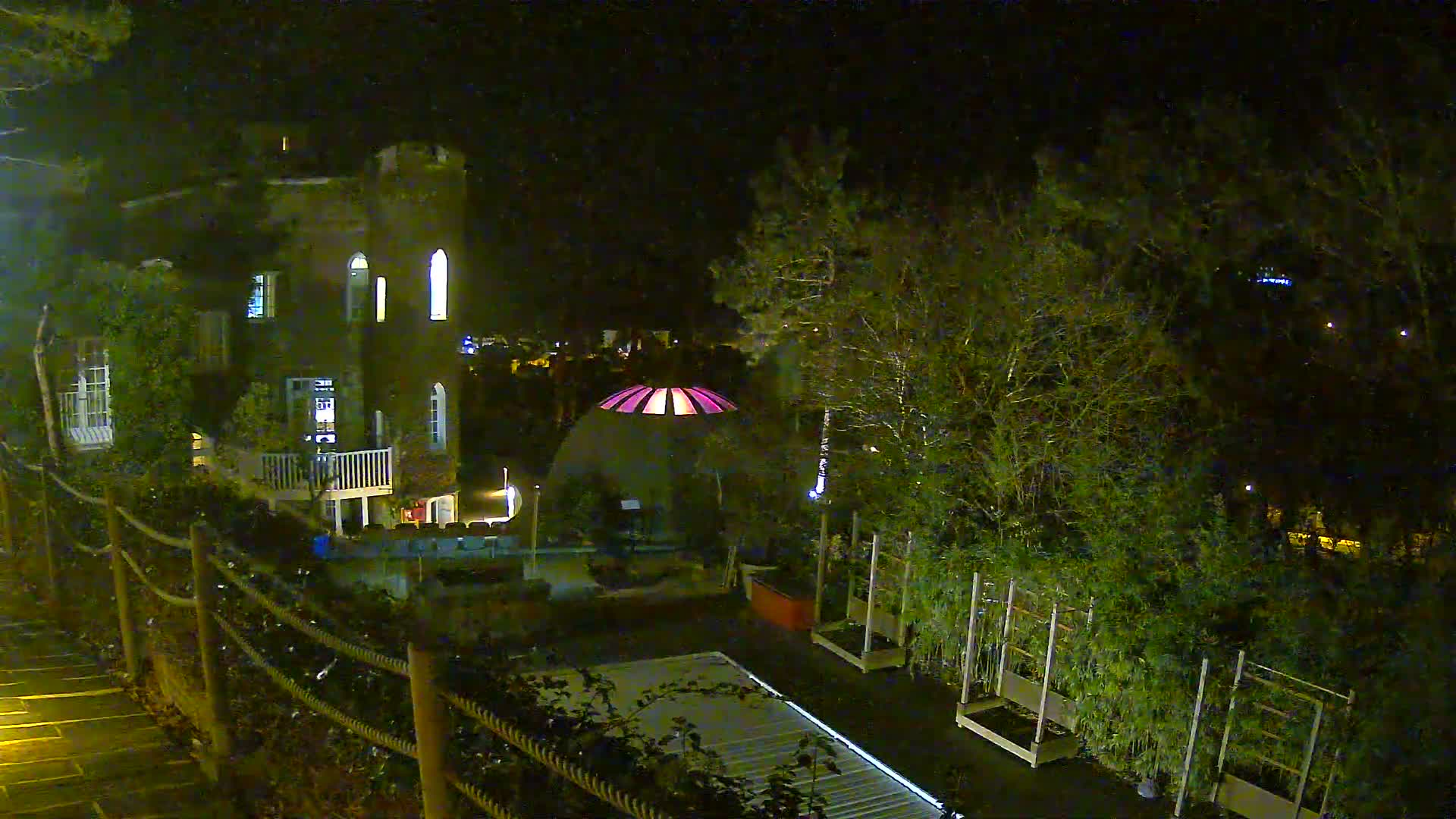An ivy-covered brick building with a conical tower overlooks a garden featuring a geodesic dome, a damp patio, and numerous trees, with a coastal town and the sea visible in the distance under a bright, partly cloudy sky.