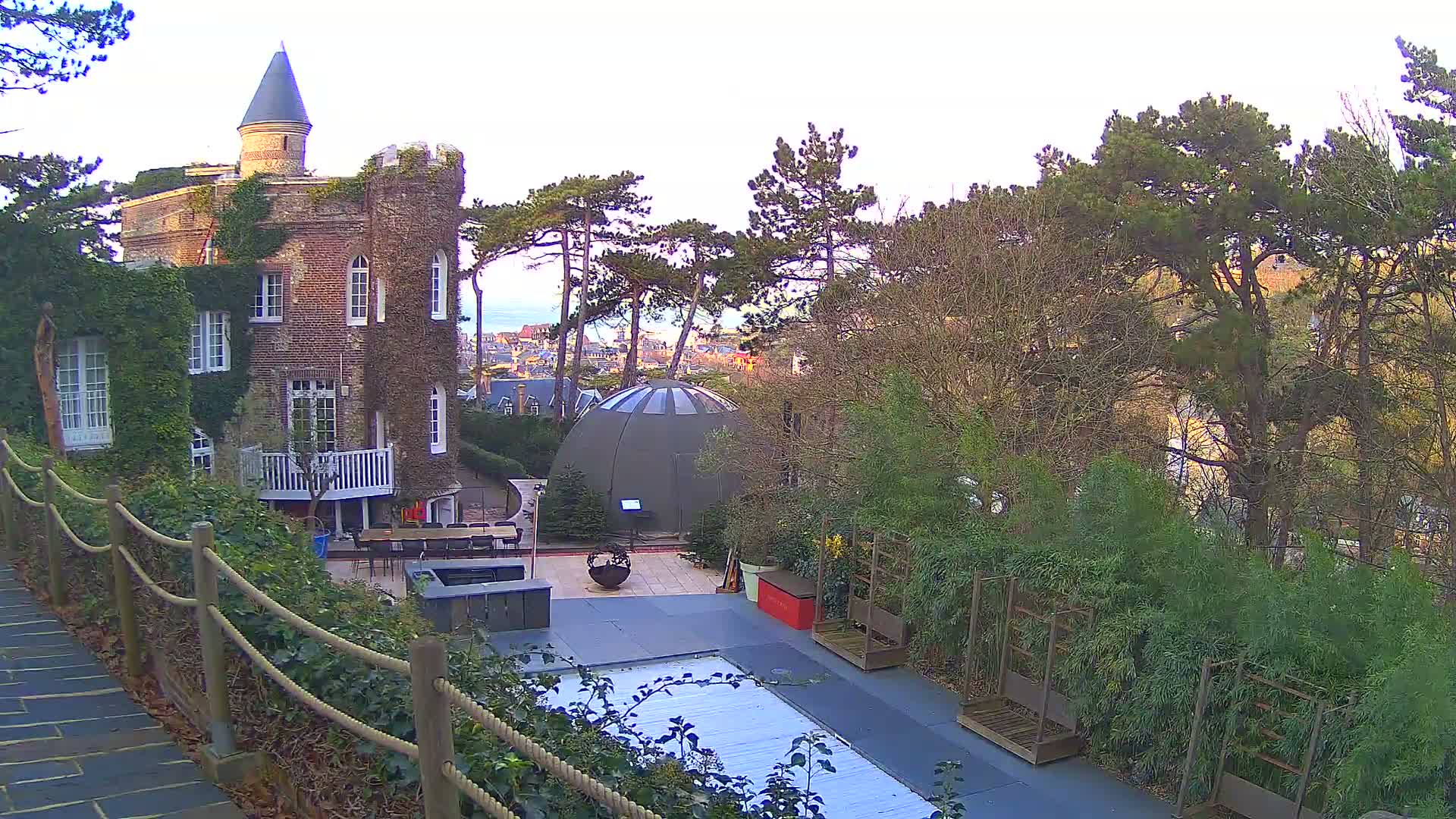 An ivy-covered brick building with a conical tower overlooks a garden featuring a geodesic dome, a damp patio, and numerous trees, with a coastal town and the sea visible in the distance under a bright, partly cloudy sky.