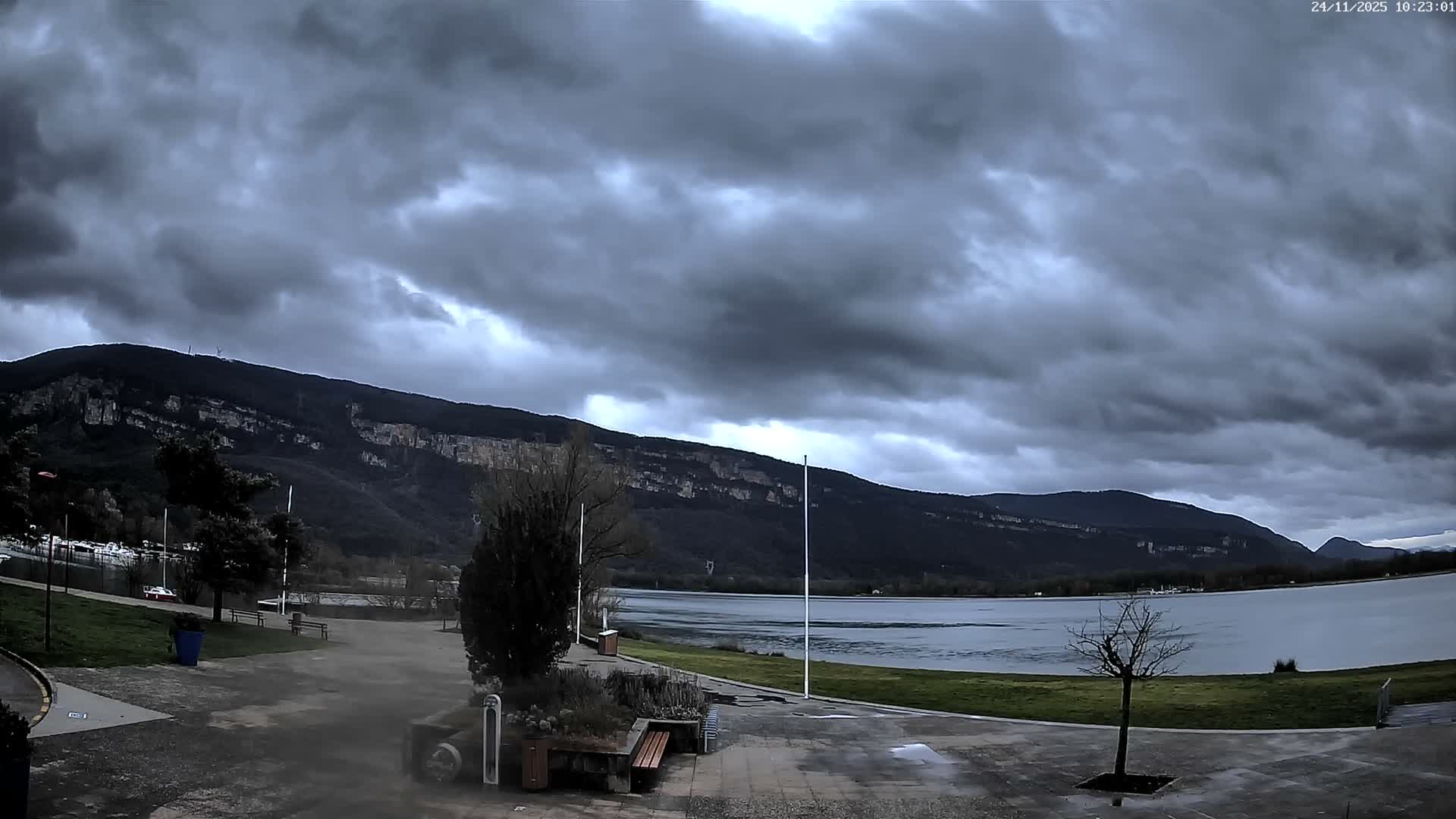 A gloomy, heavily overcast sky looms over a wide lake, bordered by dark mountains and a lakeside park with bare trees and benches in the foreground.