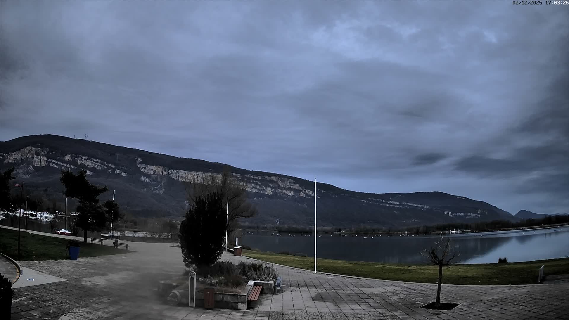 A cloudy, overcast day reveals a wide lake or river reflecting the dark sky, flanked by a paved promenade with benches and scattered trees in the foreground, and backed by a majestic range of dark mountains with visible rock faces.
