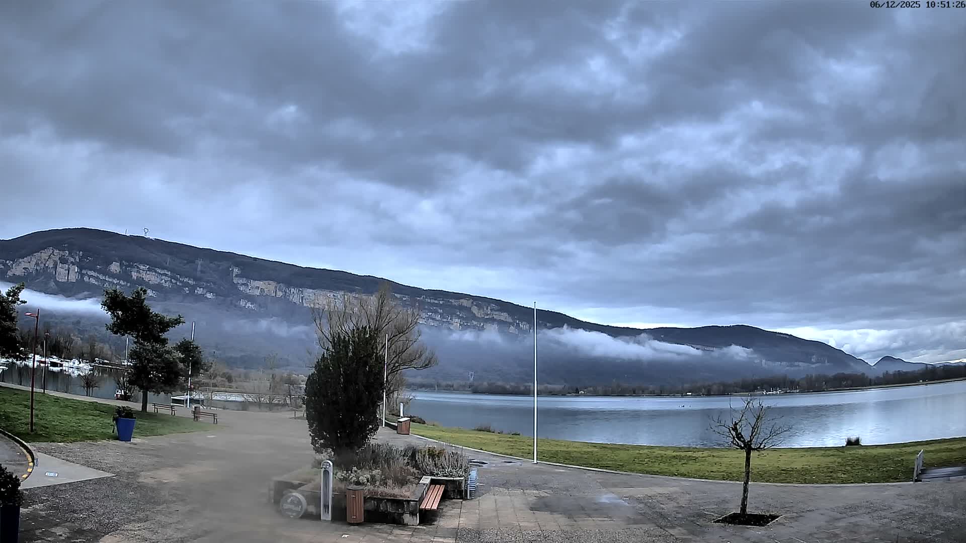 An overcast and cloudy day reveals a tranquil lake surrounded by a paved promenade and grassy banks, with distant mountains partially enshrouded in low-hanging fog and mist.