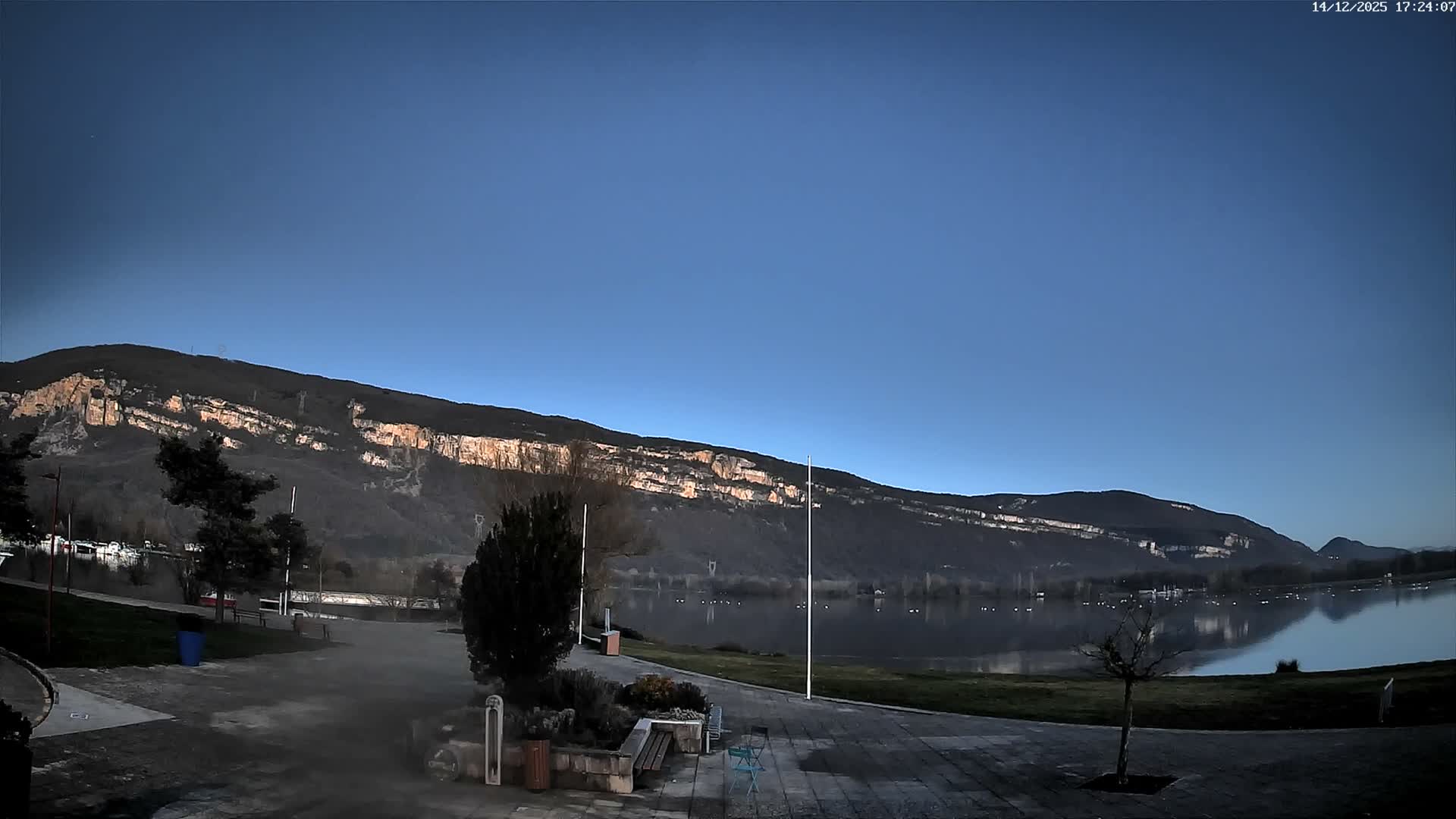 An overcast and cloudy day reveals a tranquil lake surrounded by a paved promenade and grassy banks, with distant mountains partially enshrouded in low-hanging fog and mist.