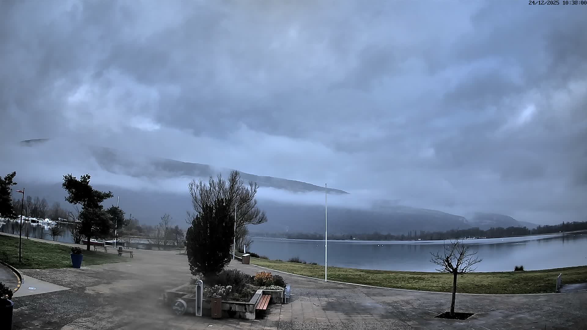 An overcast and cloudy day reveals a tranquil lake surrounded by a paved promenade and grassy banks, with distant mountains partially enshrouded in low-hanging fog and mist.