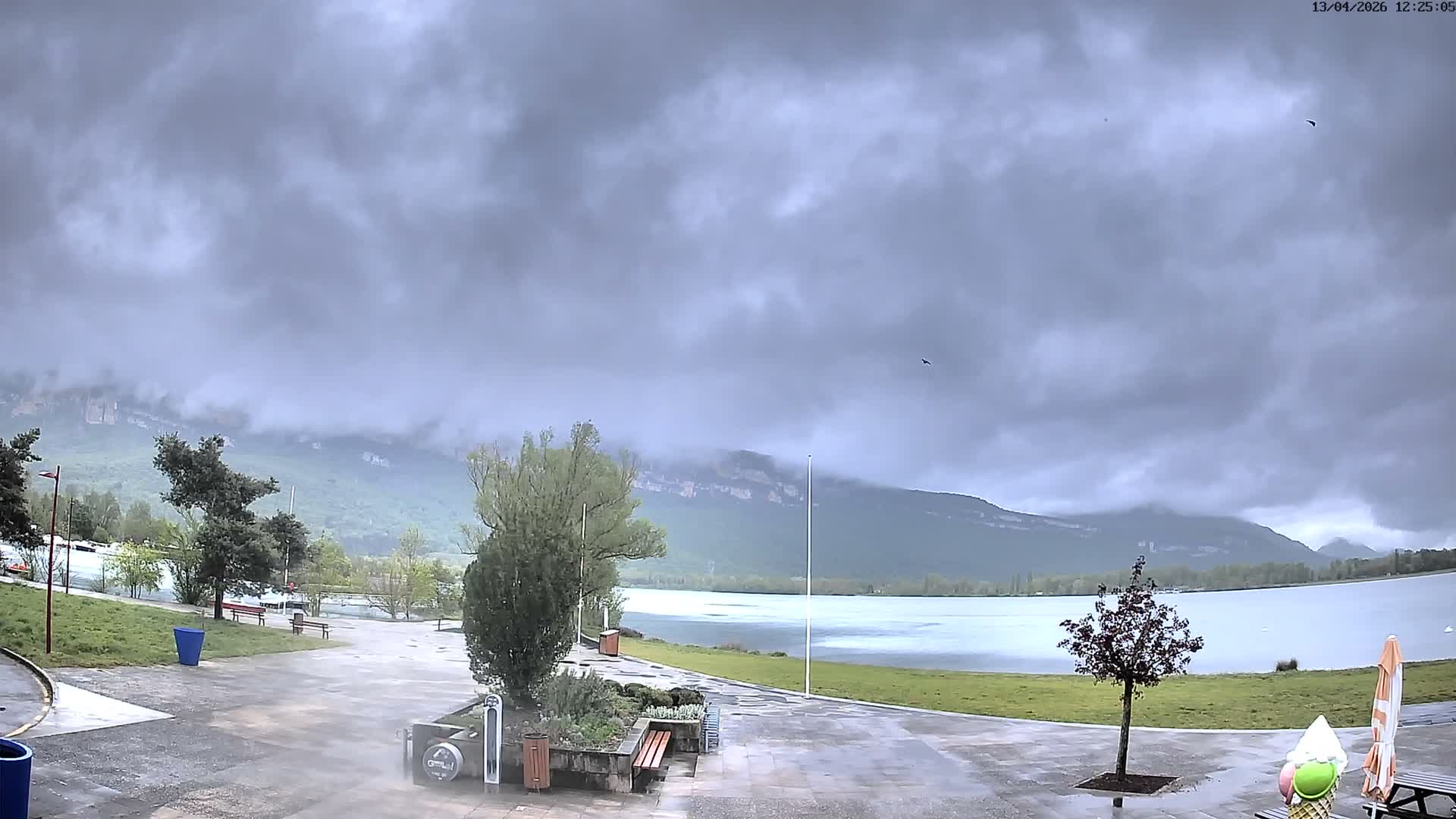 An overcast and cloudy day reveals a tranquil lake surrounded by a paved promenade and grassy banks, with distant mountains partially enshrouded in low-hanging fog and mist.