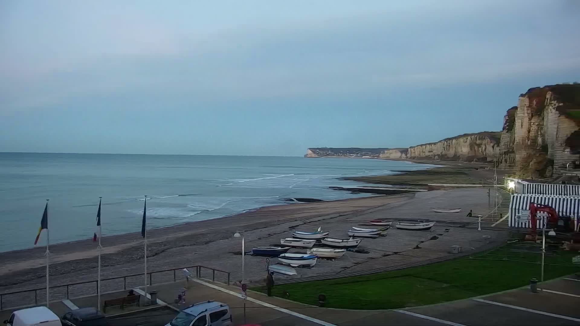A wide coastal view under an overcast sky reveals a pebble beach lined with various small boats pulled ashore, dramatic white cliffs extending into the distance, and gentle waves, with flags flying above a paved promenade in the foreground.