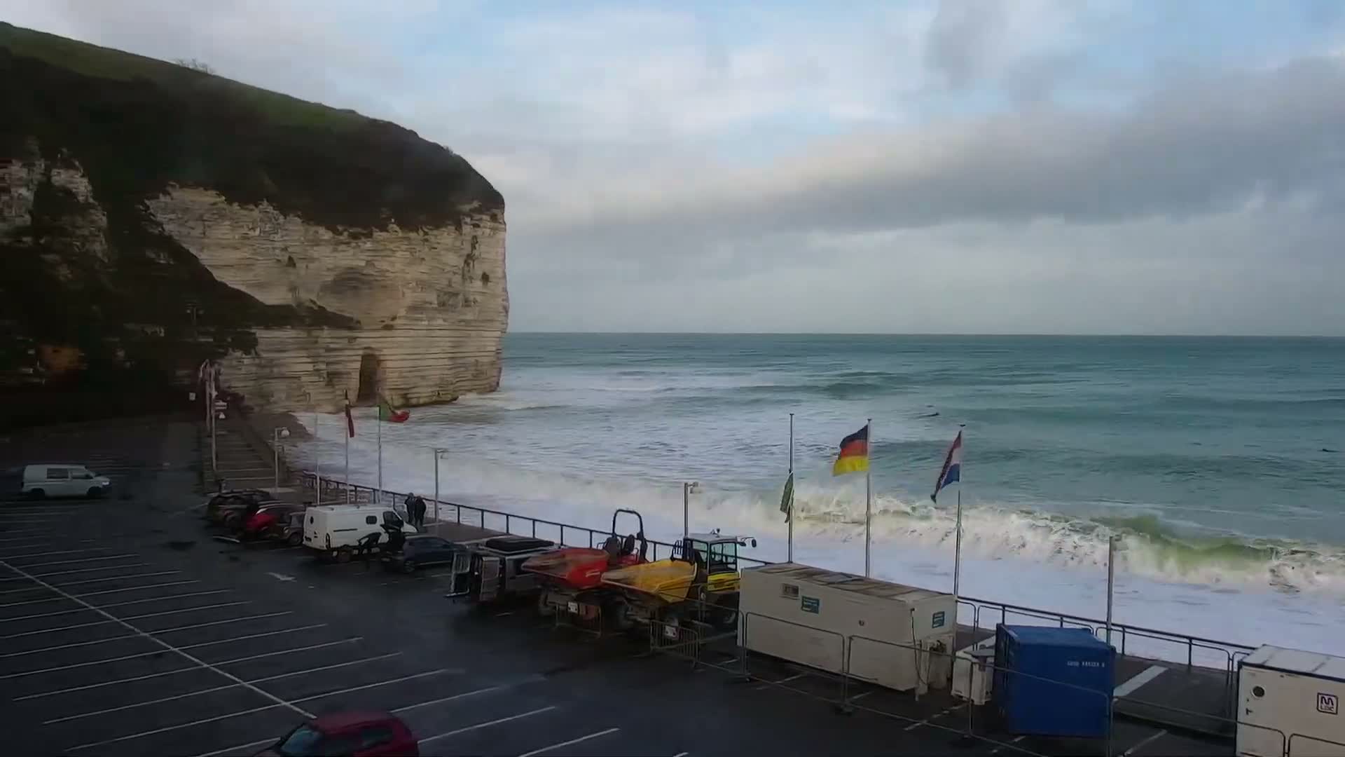 On a cloudy and breezy day, large waves crash against a coastal promenade lined with parked vehicles, utility containers, and international flags, all overlooked by a towering white chalk cliff.