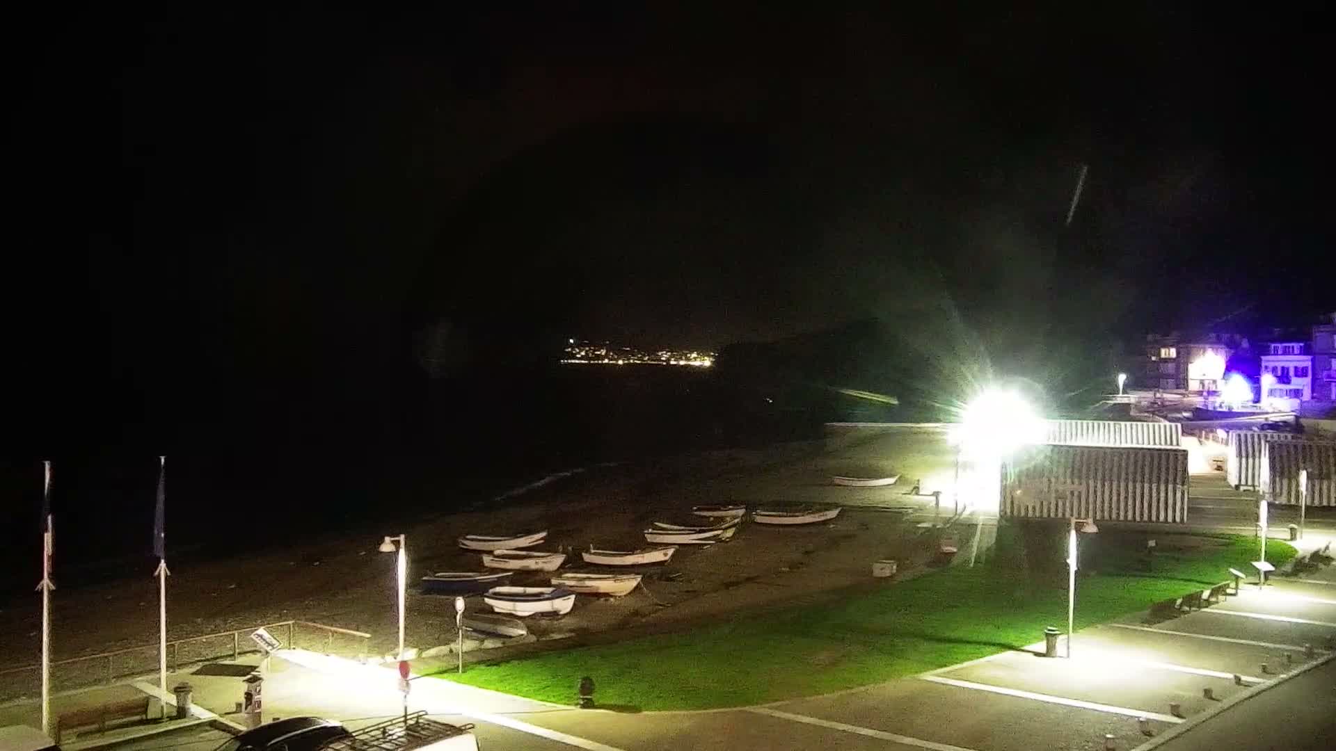On a cloudy and breezy day, large waves crash against a coastal promenade lined with parked vehicles, utility containers, and international flags, all overlooked by a towering white chalk cliff.
