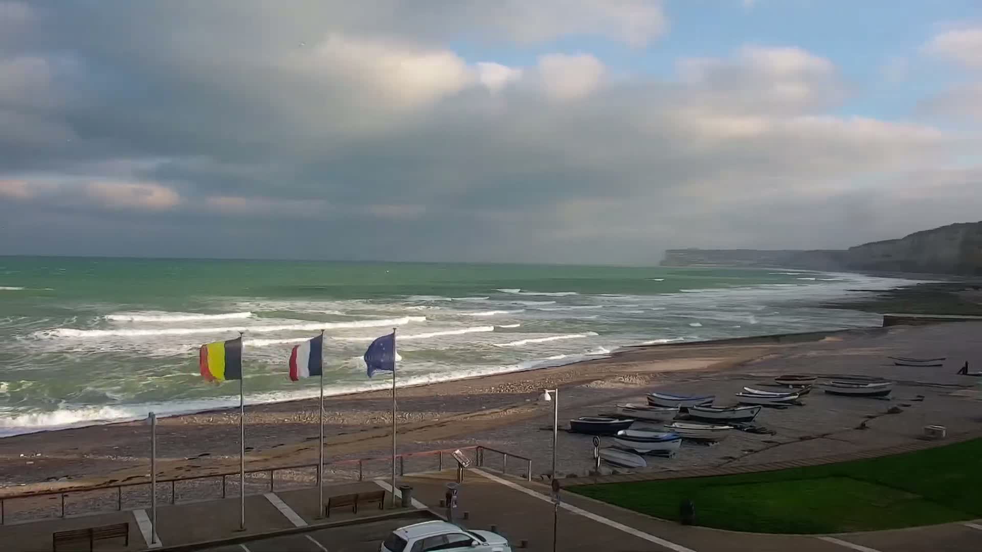 On a cloudy and breezy day, large waves crash against a coastal promenade lined with parked vehicles, utility containers, and international flags, all overlooked by a towering white chalk cliff.