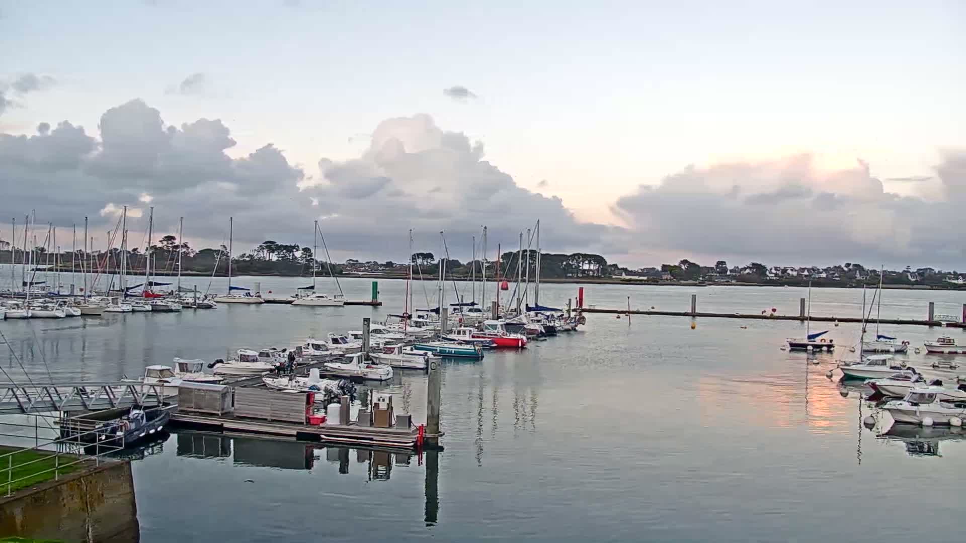 A tranquil marina is filled with numerous docked sailboats and motorboats, reflecting the calm water under a partly cloudy sky with soft, subdued light.