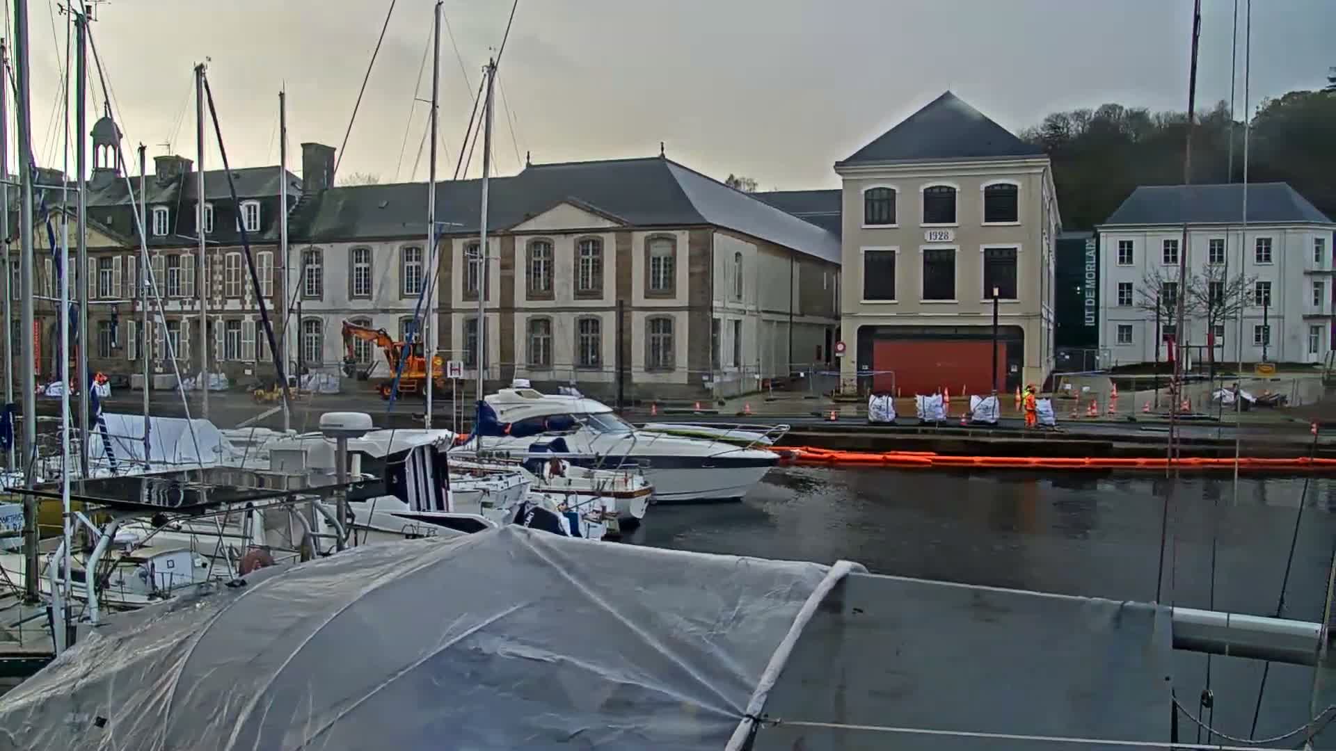A cloudy day hangs over a European harbor filled with numerous docked boats, some covered, with multi-story historic buildings lining the bustling shore where construction activity, workers, and orange water booms are visible.