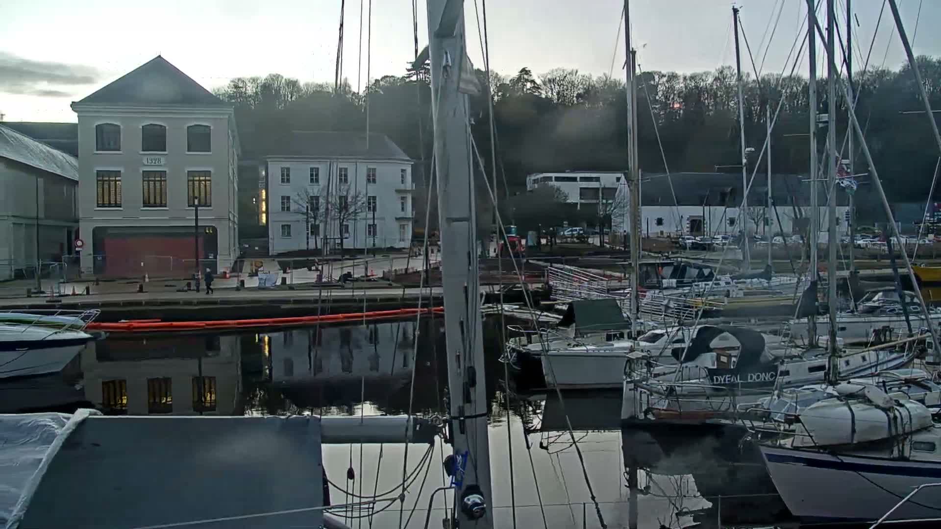A marina filled with numerous sailboats is shown under an overcast sky, with historic-looking buildings lining the waterfront and a forested hill in the background.