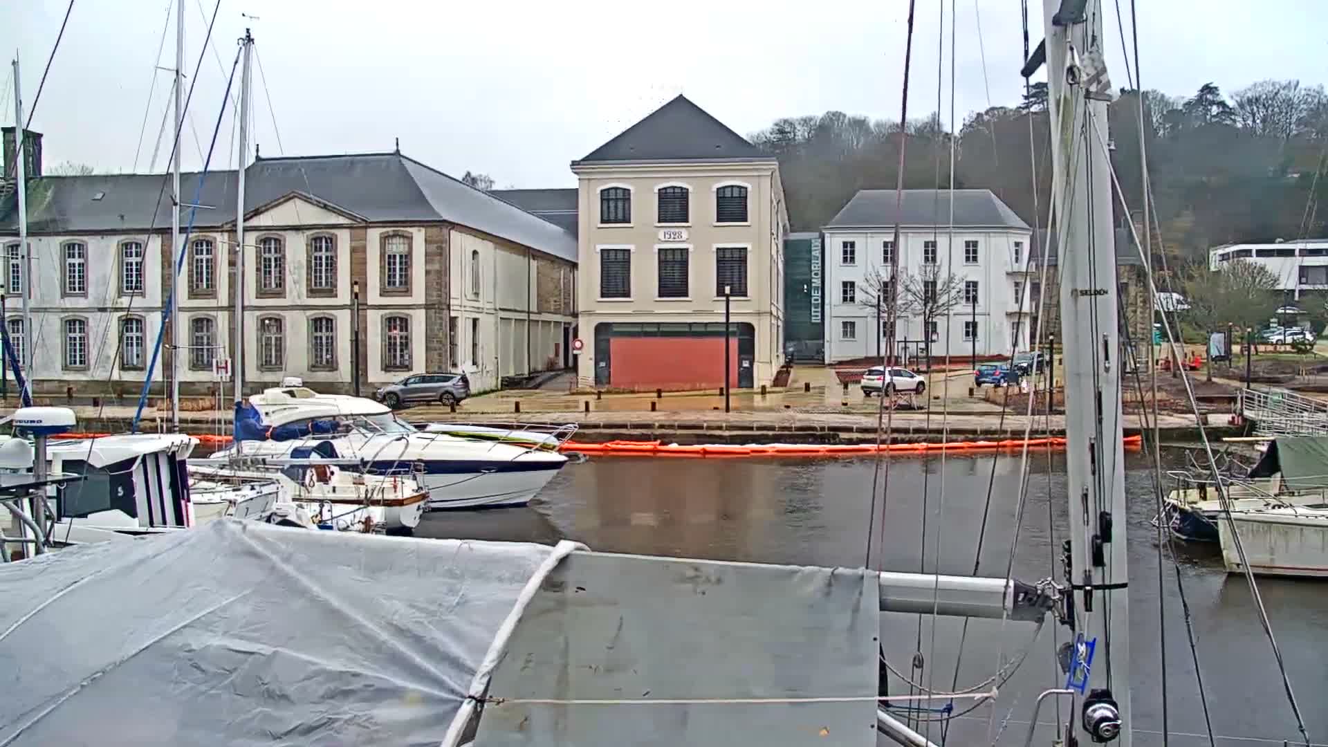 An overcast day view from a boat's rigging overlooks a marina with several boats docked, traditional buildings lining the waterfront, and a wooded hill in the background.