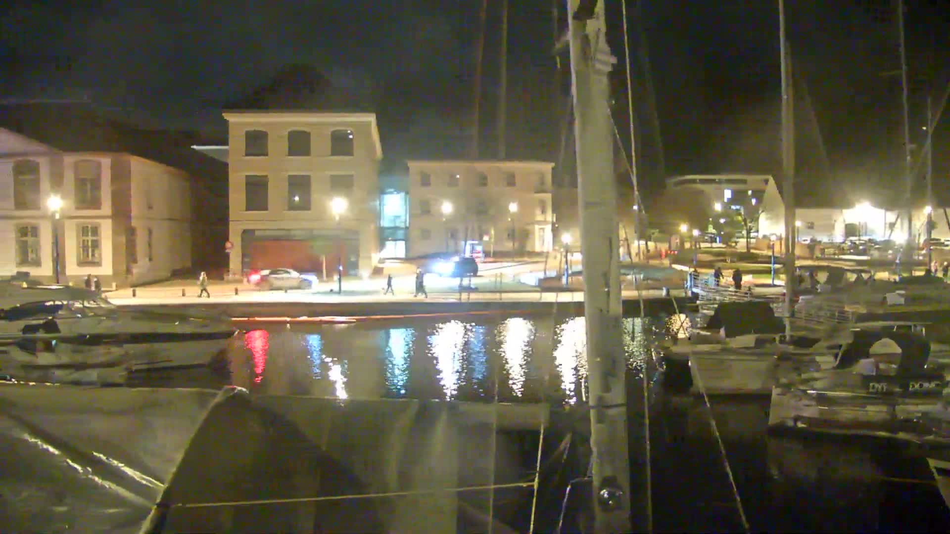 An overcast day view from a boat's rigging overlooks a marina with several boats docked, traditional buildings lining the waterfront, and a wooded hill in the background.